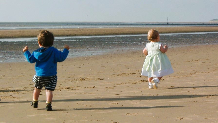 Boy toddler chasing girl toddler along a beach.